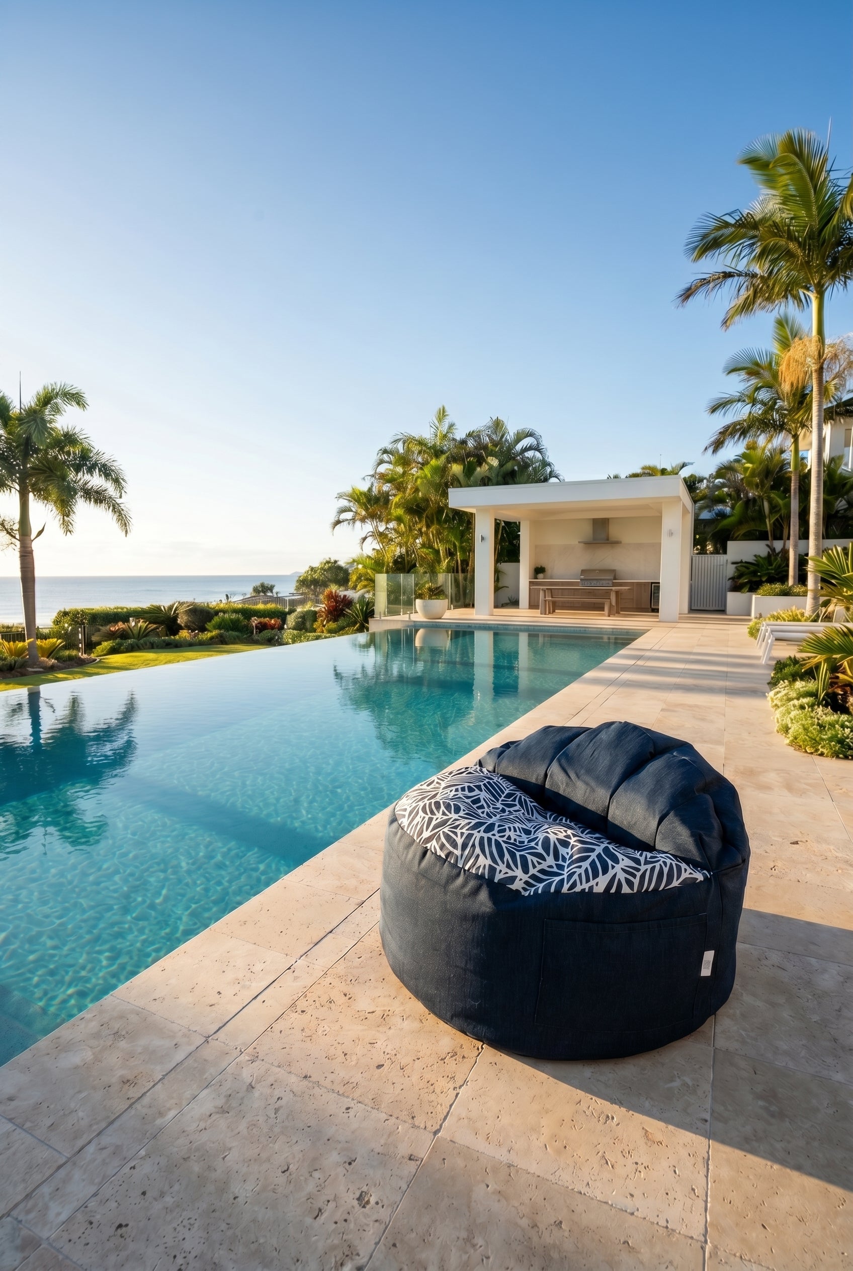 Black bean bag chair with a patterned cushion by a poolside with palm trees and a clear sky.