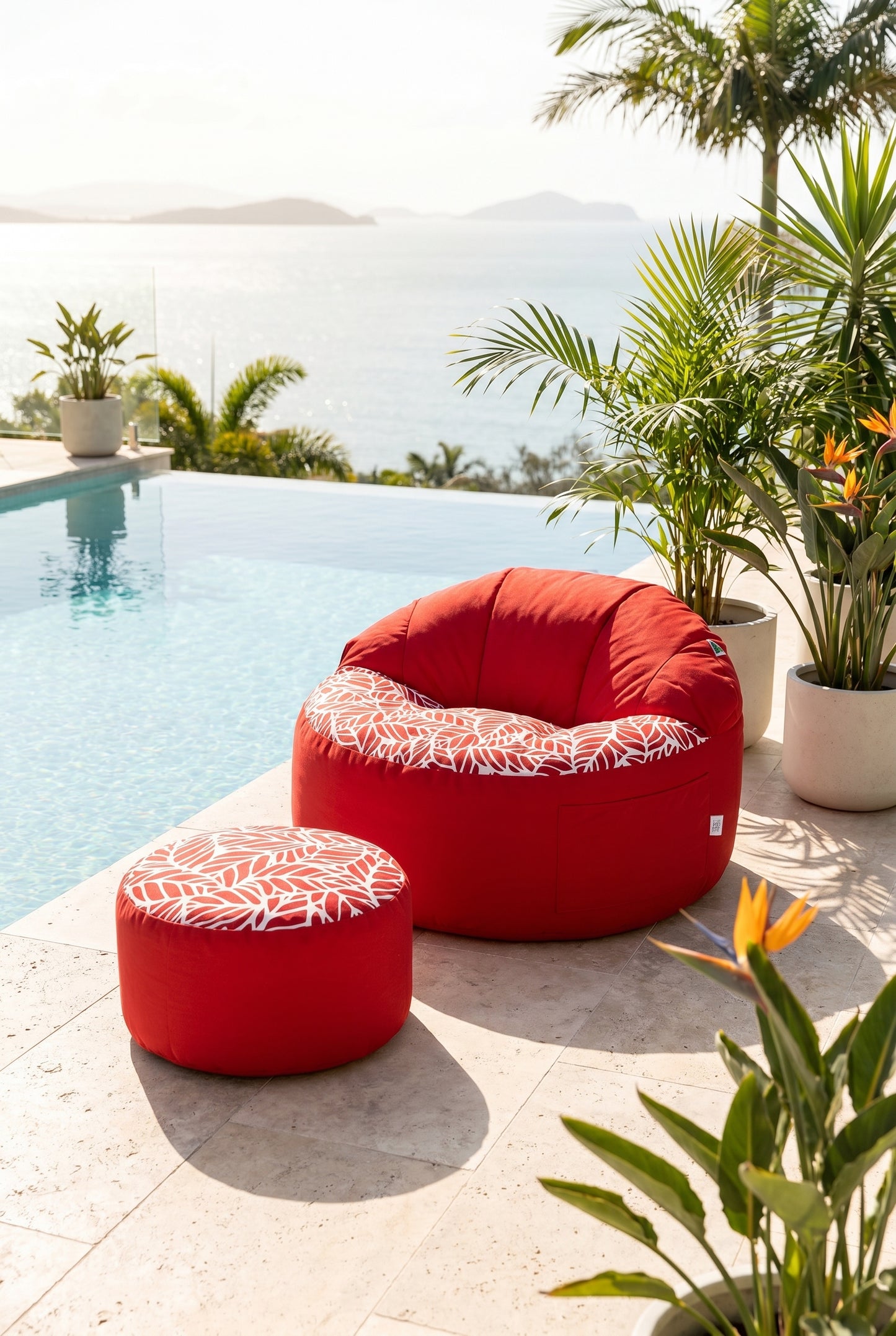 Red outdoor bean bags with white patterns by a poolside with plants and mountains in the background