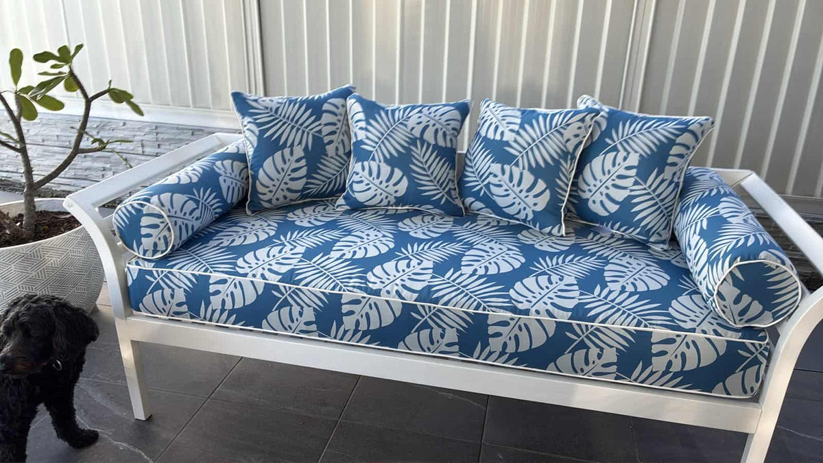 A white outdoor patio sofa with blue and white tropical monstera leaf patterned cushions, shown on a tiled patio with a potted plant and a small black dog nearby. 